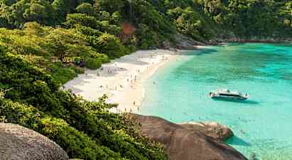 Hiekkaranta ihmisten kanssa, vehreiden puiden ympäröimä ja kirkkaan turkoosin veden reunustama. Vene on lähellä rantaa. (Tekoälyn luoma teksti)