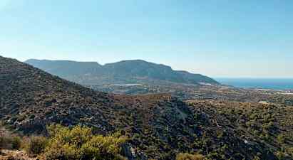 Maisema mäkisestä maastosta, jossa on vaihtelevaa kasvillisuutta, ja taustalla näkyy meri kirkkaan sinisen taivaan alla. (Tekoälyn luoma teksti)