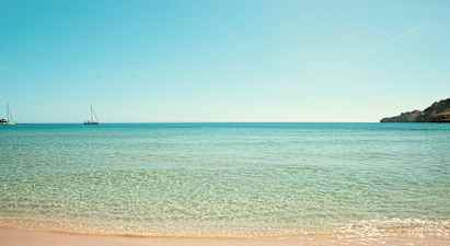 Ranta, jossa kirkasta vettä ja kaksi purjevenettä merellä. (Tekoälyn luoma teksti)