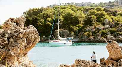 Purjevene on kiinnitetty rauhalliseen poukamaan kallioiden ja metsän ympäröimänä, rannalta kalastavan henkilön kanssa. (Tekoälyn luoma teksti)