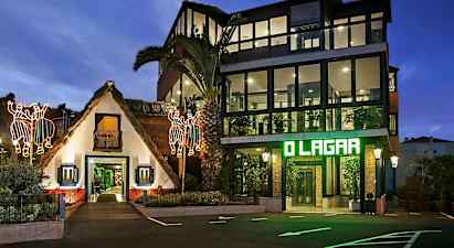 Rakennus, jossa on perinteinen olkikatto ja nykyaikainen lasipaneelirakennus sen vieressä, valaistuna iltaisin hohtavilla koristeilla. (Tekoälyn luoma teksti)