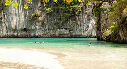 Kuvassa on ranta, jonka kirkkaan turkoosi vesi on korkeiden kallioiden ympäröimä ja kasvien peittämä. Useita henkilöitä ui vedessä kirkkaan taivaan alla. (Tekoälyn luoma teksti)