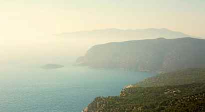 Rannikko, jossa on jyrkkiä kallioita ja vihreitä metsiä, jotka laskeutuvat mereen. Maisemaa peittää utu. (Tekoälyn luoma teksti)