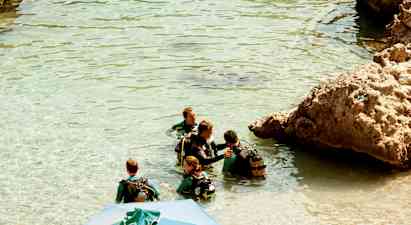 Ryhmä ihmisiä märkäpuvuissa ja sukellusvarusteissa seisoo kirkkaassa vedessä kallioiden vieressä. (Tekoälyn luoma teksti)