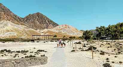 Kuiva maisema, jossa ihmisiä kävelee polkua pitkin kohti pieniä rakennuksia. Vuori näkyy taustalla kirkkaan sinisen taivaan alla. (Tekoälyn luoma teksti)