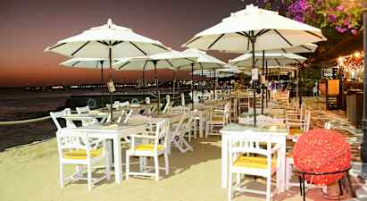 Valkoiseksi maalattuja pöytiä ja tuoleja suurilla varjoilla rannalla hämärän aikaan. (Tekoälyn luoma teksti)