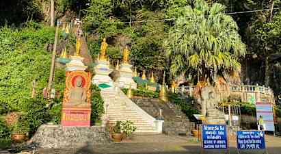 Kuva portaat, jotka johtavat useisiin kultaisiin buddhalaisiin patsaisiin ja rakenteisiin vehreyden ympäröimänä. (Tekoälyn luoma teksti)