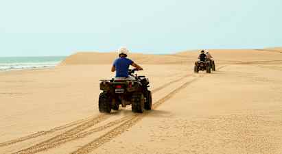 Kaksi henkilöä ajaa mönkijöillä hiekkarannalla, meri taustalla. (Tekoälyn luoma teksti)