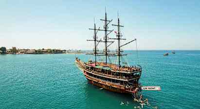 Suuri purjevene puusta, joka kelluu rauhallisilla vesillä uimareiden ympäröimänä, taustalla rannikkoa. (Tekoälyn luoma teksti)