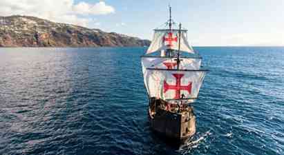 Purjelaiva, jonka suuret valkoiset purjeet on koristeltu punaisilla risteillä, purjehtii sinisen meren yllä lähellä mäkistä rannikkoa. (Tekoälyn luoma teksti)