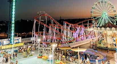 Kuvassa on vilkas huvipuisto illalla, jossa on suuri maailmanpyörä, värikäs vuoristorata ja allas törmäilyautoilla. (Tekoälyn luoma teksti)