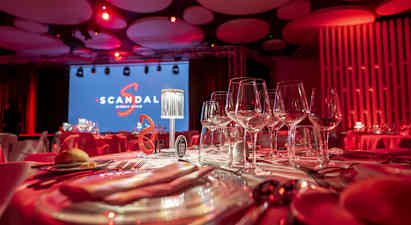 Pöydän kattaus viinilaseilla huoneessa, jossa on punainen valaistus. Taustalla suuri näyttö, jossa teksti 'Scandal', sekä lava. (Tekoälyn luoma teksti)