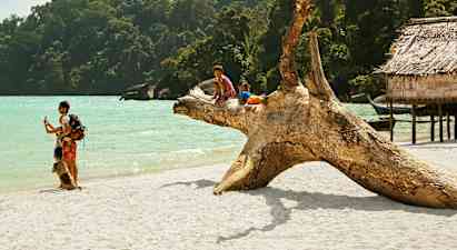 Kaksi lasta istuu suurella puunrungolla hiekkarannalla meren äärellä, ympärillään vehreää metsää ja puu-rakennuksia. (Tekoälyn luoma teksti)