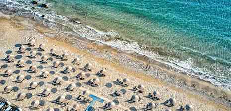 Ilmakuva hiekkarannasta, jossa aurinkotuoleja ja aurinkovarjoja, taustalla rantaviiva ja sininen meri. (Tekoälyn luoma teksti)