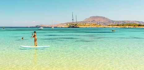 Henkilö seisoo sup-laudalla kirkkaassa sinisessä vedessä, taustalla vuoristoinen rantaviiva. (Tekoälyn luoma teksti)