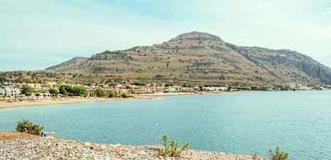 Rannikko kivirannalla ja kirkkaalla vedellä, taustalla näkyy taloja ja korkea mäki. (Tekoälyn luoma teksti)