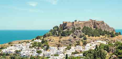 Näkymä kukkulalle, jolla historiallinen linnoitus, ympäröivänä vihreitä puita ja valkoisia rakennuksia lähellä merta. (Tekoälyn luoma teksti)