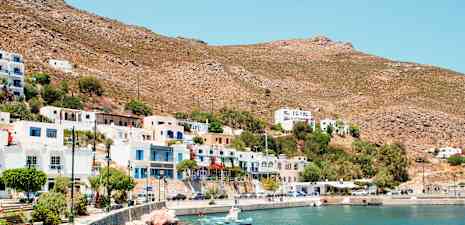 Rannikolla sijaitseva kaupunki, jossa on valkoisia ja värikkäitä rakennuksia, pieniä veneitä kivisen rannikon äärellä ja kuivaa vuoristoa taustalla. (Tekoälyn luoma teksti)