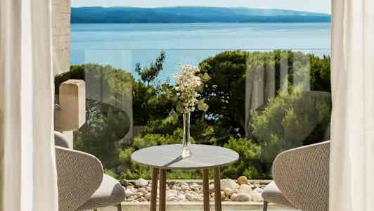 Pöytäkoriste, jossa kukkia, pöydän ääressä ikkunan edessä, näkymä merelle ja vehreä luonto. (Tekoälyn luoma teksti)