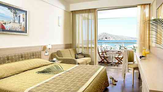 Hotellihuone, jossa sänky, istuinalue ja näkymä merelle parvekkeen kautta, jossa on tuoleja ja pöytä. (Tekoälyn luoma teksti)