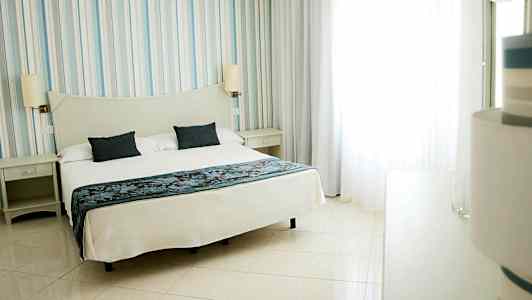 Hotellihuone, jossa on suuri sänky, valkoisilla vuodevaatteilla ja kaksi tummansinistä tyynyä. (Tekoälyn luoma teksti)