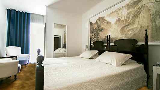 Vuode, jossa on veistetty pääty huoneessa, jossa on seinäpeili ja istumapaikkoja. (Tekoälyn luoma teksti)