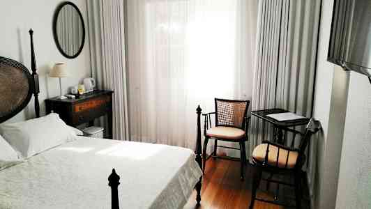 Hotellihuone, jossa on sänky, työpöytä ja tuoli sekä ikkuna valkoisilla verhoilla. (Tekoälyn luoma teksti)