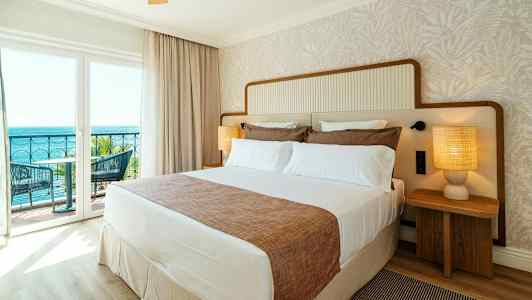 Hotellihuone, jossa on iso sänky, kaksi yöpöytää lampuilla ja parveke merinäköalalla. (Tekoälyn luoma teksti)