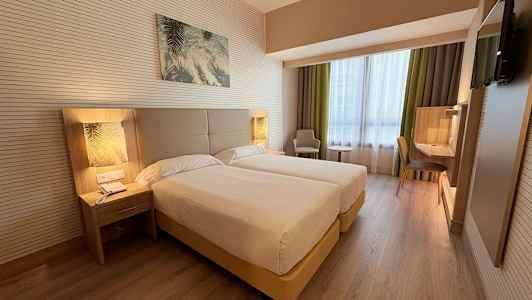 Hotellihuone, jossa on kaksi yhden hengen vuodetta, yöpöytä, työtila ja nojatuoli ikkunan vieressä. (Tekoälyn luoma teksti)