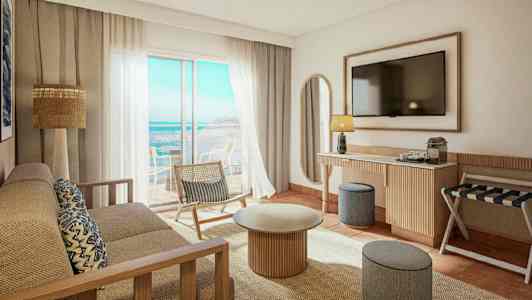 Valoisa ja moderni hotellihuone, josta on näkymä merelle. Huoneessa on sohva, sohvapöytä, tuoli ja seinälle asennettu televisio. (Tekoälyn luoma teksti)