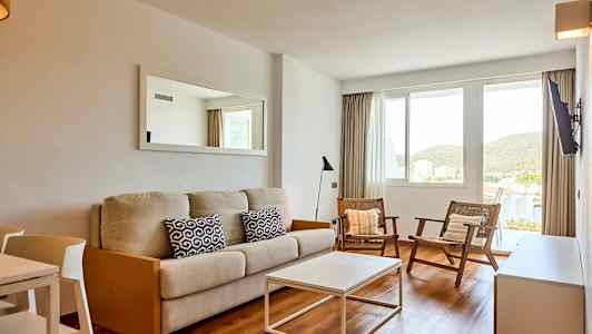 Olohuone, jossa on beige sohva, kaksi tuolia ja valkoinen sohvapöytä. Ikkunasta näkyvät vehreät maisemat. (Tekoälyn luoma teksti)