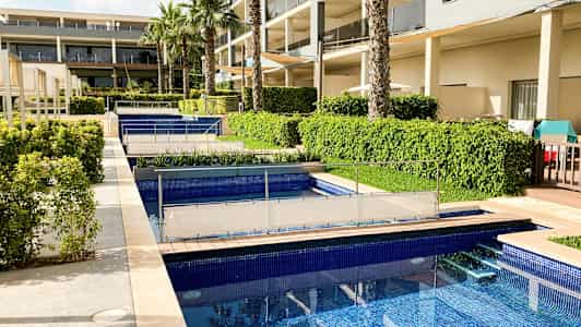 Kuva hotellialueesta, jossa on useita altaita, vihreitä pensaita ja palmuja rakennuksen lähellä. (Tekoälyn luoma teksti)