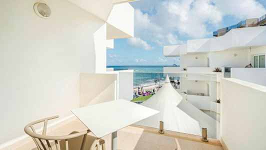 Parveke, jossa on pöytä ja tuolit, näkymä merelle ja valkoisille rakennuksille. (Tekoälyn luoma teksti)