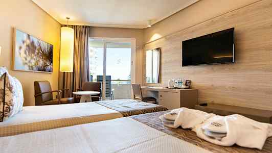 Hotellihuone, jossa on kaksi yhden hengen vuodetta, istuimia pyöreän pöydän ympärillä sekä työpiste seinään kiinnitetyllä televisiolla. (Tekoälyn luoma teksti)