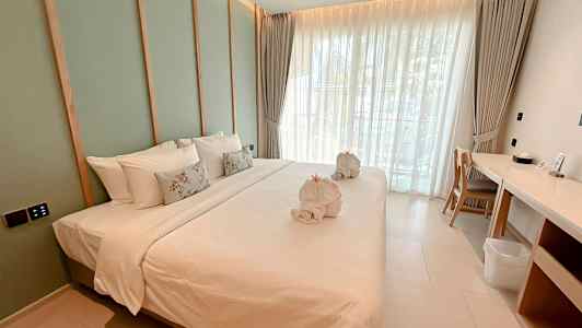 Kuva modernista hotellihuoneesta, jossa on suuri pedattu sänky, valkoiset vuodevaatteet ja koristeellisia tyynyjä sekä pyyhkeistä muotoillut joutsenet. Huoneessa on ikkunat vaaleilla verhoilla ja sivupöytä. (Tekoälyn luoma teksti)