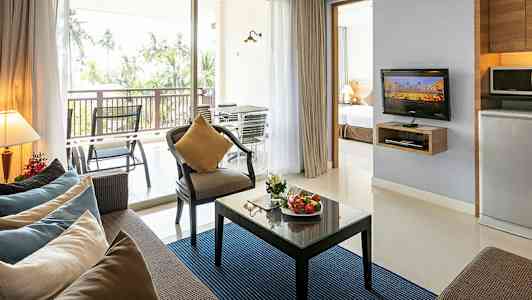 Modernisti sisustettu olohuone, jossa sohva, nojatuoli, lasipöytä hedelmillä, televisio ja keittiöosa sekä parveke näkymällä palmuihin. (Tekoälyn luoma teksti)