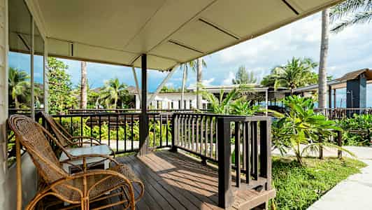 Veranta, jossa on puulattia, rottinkituolit ja näkymä vehreään ympäristöön sekä lähellä sijaitseviin rakennuksiin ja palmuihin. (Tekoälyn luoma teksti)