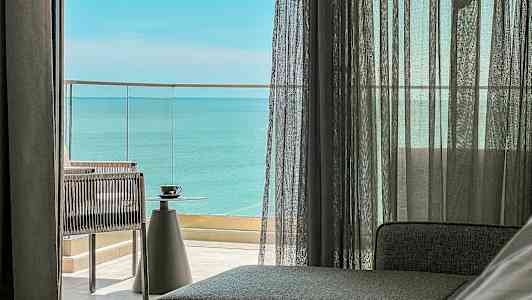 Näkymä huoneesta lasiovien kautta parvekkeelle, josta avautuu merinäköala. Pöydällä on kuppi. (Tekoälyn luoma teksti)