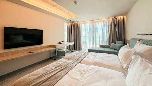 Moderni hotellihuone, jossa on parivuode, seinälle kiinnitetty televisio, työpöytä ja istuma-alue suurten ikkunoiden vieressä. (Tekoälyn luoma teksti)