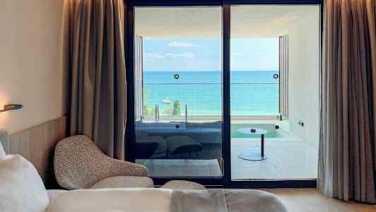 Hotellihuone, jossa on sänky ja nojatuoli, sekä näkymä parvekkeelle ja taustalla olevaan mereen. (Tekoälyn luoma teksti)