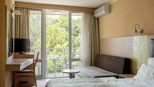 Hotellihuone, jossa on sänky, kirjoituspöytä, tuoli ja sohva. Ikkuna ja parveke, josta on näkymä vehreyteen. (Tekoälyn luoma teksti)