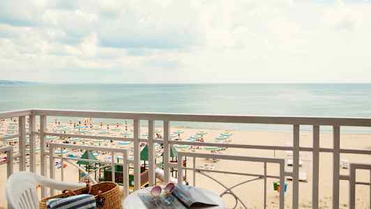 Näkymä parvekkeelta rannalle, jolla on aurinkovarjoja ja aurinkotuoleja, sekä taustalla oleva meri. (Tekoälyn luoma teksti)