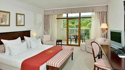 Hotellihuone, jossa vuode, istuinryhmä ja parveke näkymällä vehreään maisemaan. (Tekoälyn luoma teksti)