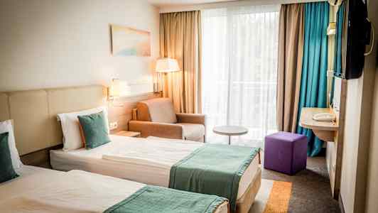 Kaksi yhden hengen vuodetta turkooseilla päiväpeitteillä hotellihuoneessa, jossa on ikkuna ja istuinryhmä. (Tekoälyn luoma teksti)