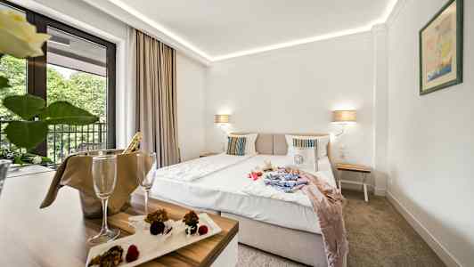 Hotellihuone, jossa on parivuode, yöpöytä ja parveke näkymä puiden suuntaan. Pöydällä samppanjajäähdytin ja jälkiruoka. (Tekoälyn luoma teksti)