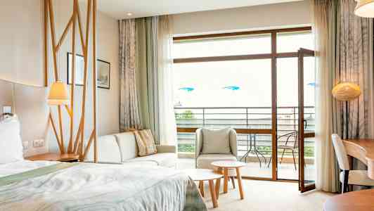 Hotellihuone, jossa on sänky, oleskelualue, pöytä ja avoin parvekkeen ovi. (Tekoälyn luoma teksti)