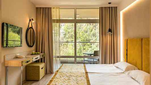 Hotellihuone, jossa on parisänky, keltainen sängynpääty, pieni kirjoituspöytä ja suuri ikkunaseinä kohti luontoa. (Tekoälyn luoma teksti)