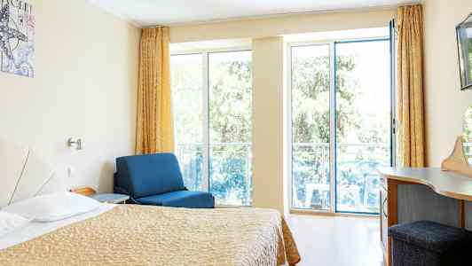 Huone, jossa sänky, sininen nojatuoli ja kirjoituspöytä, suuret ikkunat parvekkeelle. (Tekoälyn luoma teksti)