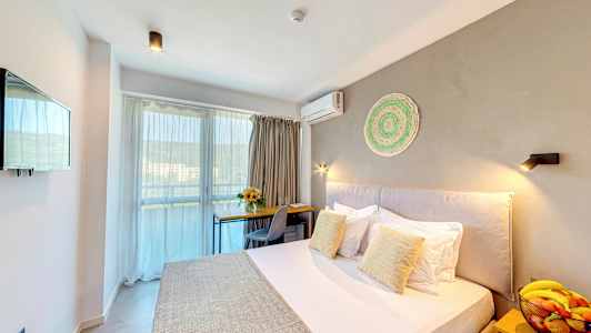 Hotellihuone, jossa on sänky, kirjoituspöytä, tuoli, kulho hedelmiä yöpöydällä ja parveke taustalla. (Tekoälyn luoma teksti)