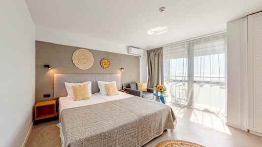 Vaalea hotellihuone, jossa suuri sänky, harmaa sohva ja parveke näköalalla. (Tekoälyn luoma teksti)